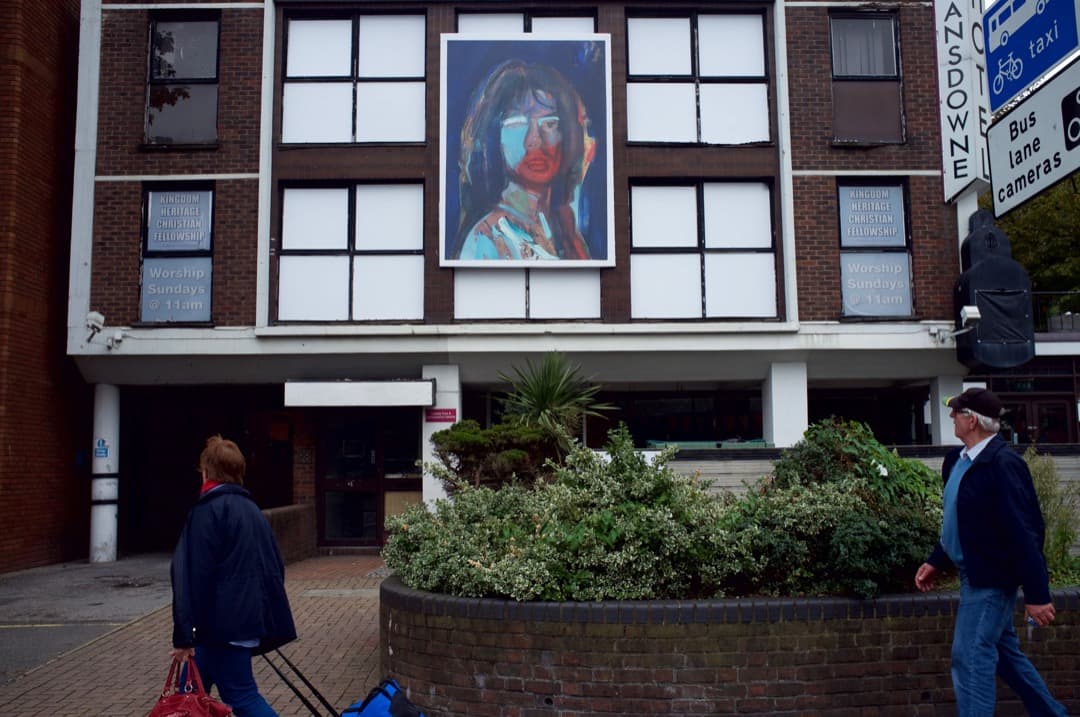 Photo of a women with a bag and a man looking at artwork on the side of a 1960s church building